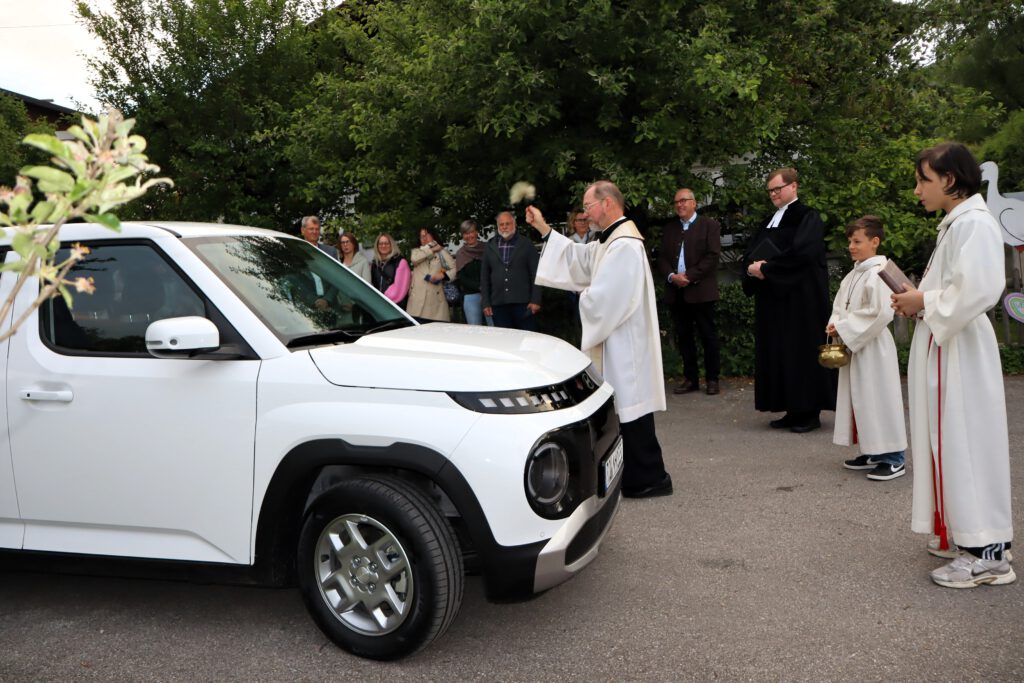Bei einer kirchlichen Fahrzeugsegnung sprengt ein Priester Weihwasser auf einen weißen Geländewagen, daneben stehen Messdiener und Gemeindemitglieder.