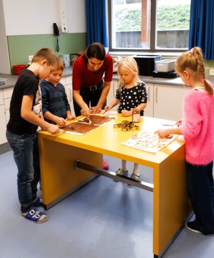 Kinder backen gemeinsam mit einer Betreuerin am gelben Tisch in der Mittags- und Hausaufgabenbetreuung der Leo-von-Welden-Schule Bad Feilnbach.